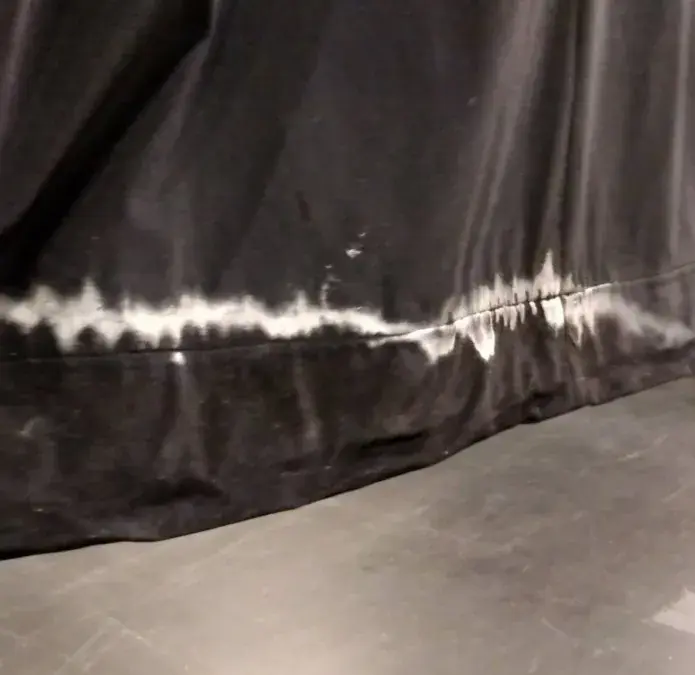 Water staining and a salt-tide line wicking up from the bottom hem of a stage drape after moisture exposure.