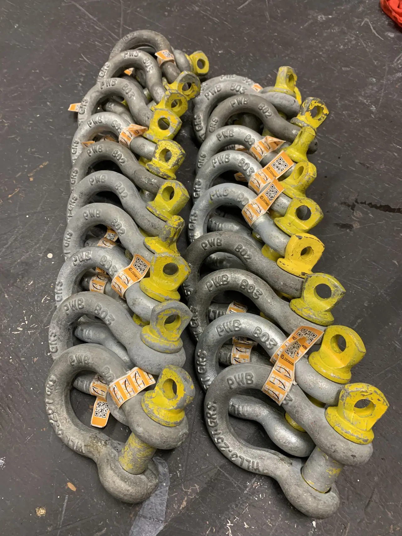 Two rows of grey bow shackles with yellow pins and asset tags laid out on a workshop floor.