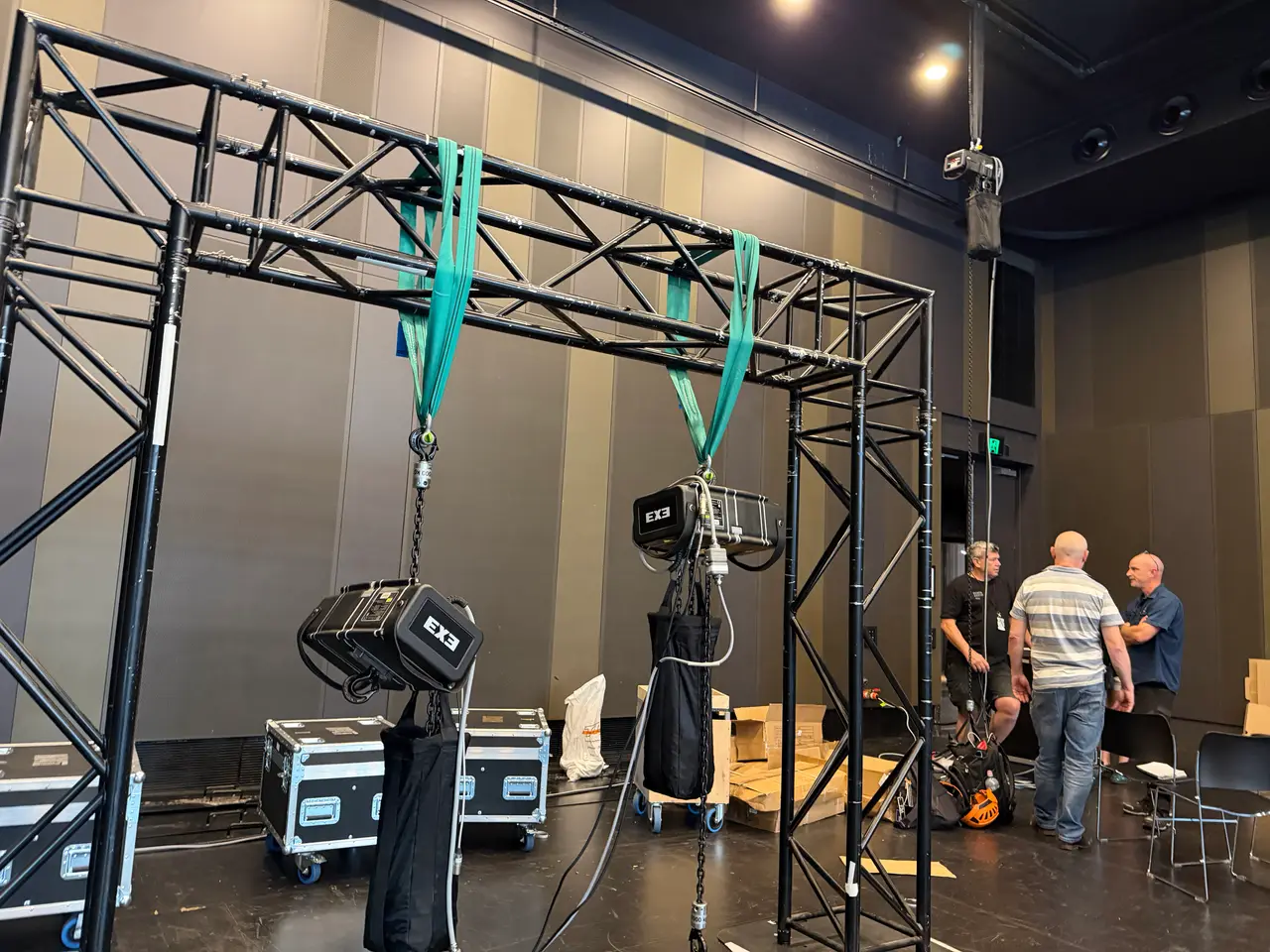 Two EXE chain hoists hang from a black truss frame in a theatre space while crew talk behind them.