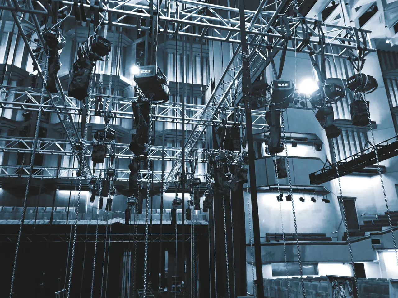 A fly tower rigging grid of chain hoists and truss above rows of auditorium seating.