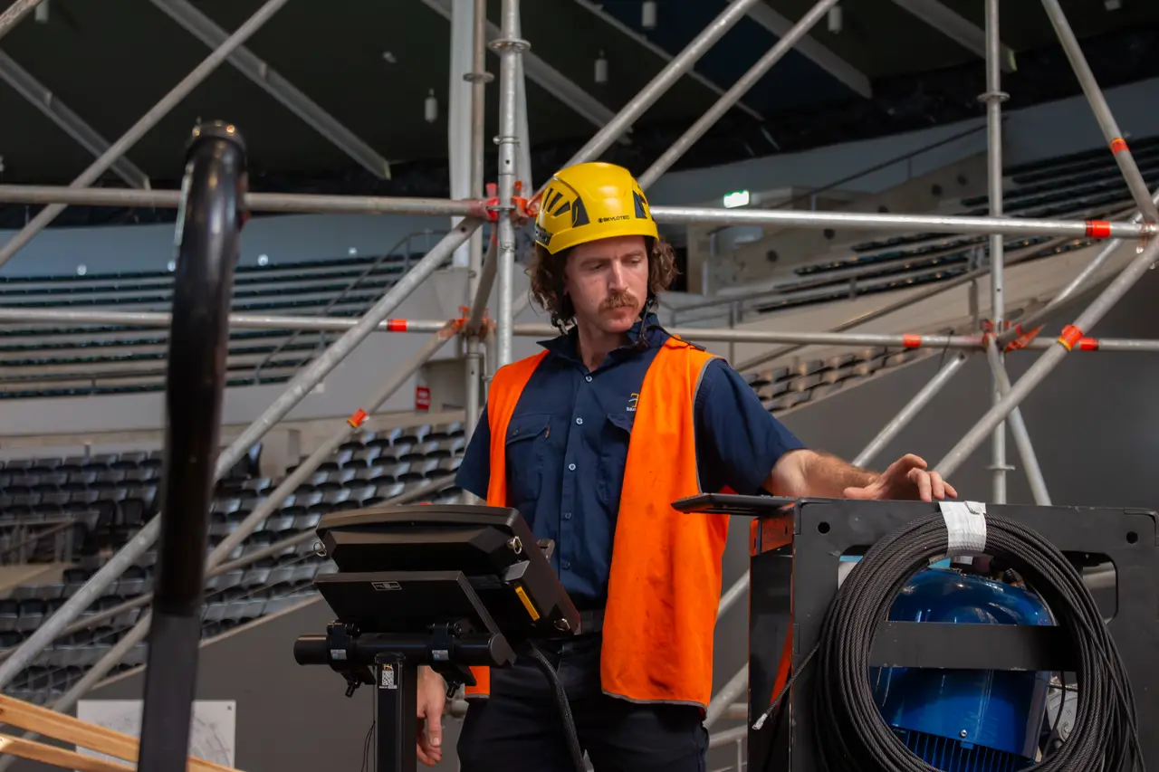 A TSA crew member in hi-vis and a hard hat works at a hoist control console inside an arena.