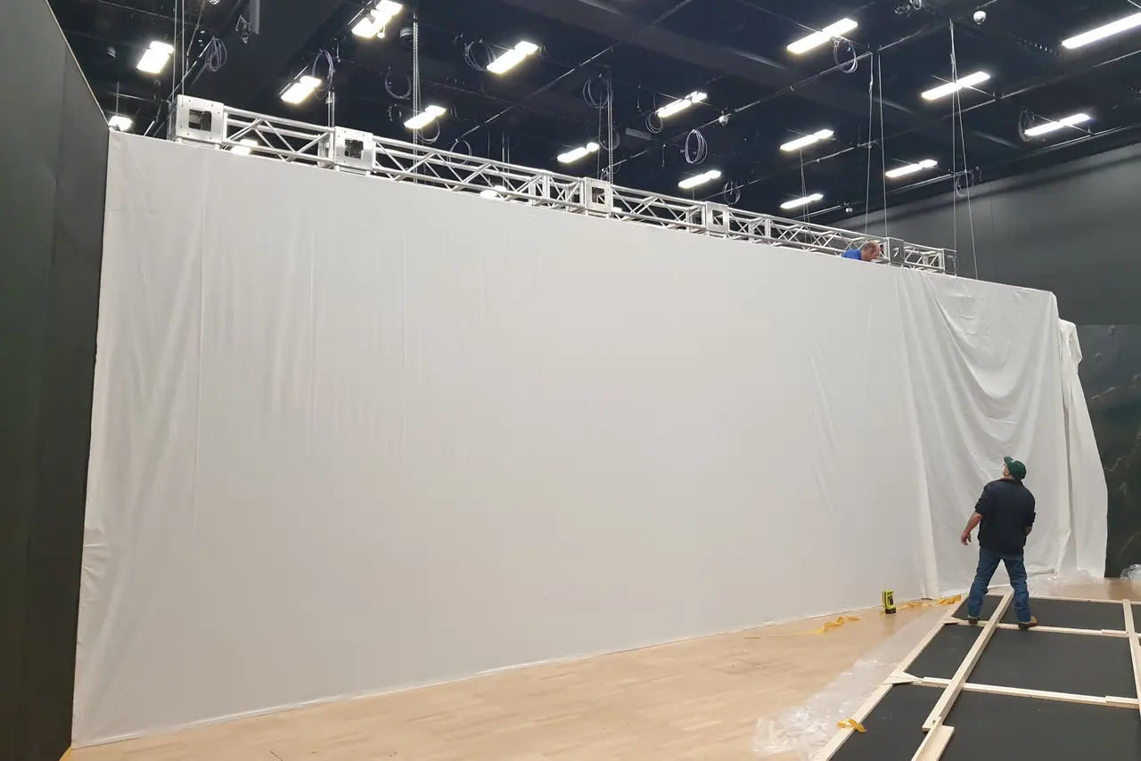 Installers rigging a tall white cyclorama wall under a truss in an exhibition space