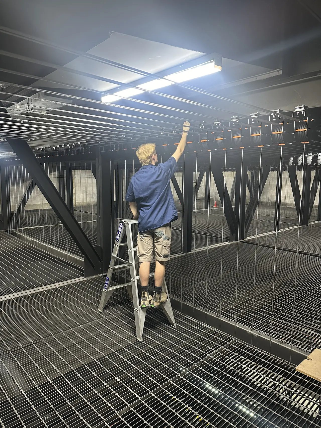 A technician on a step ladder checks a row of loft block pulleys over a steel mesh fly floor.