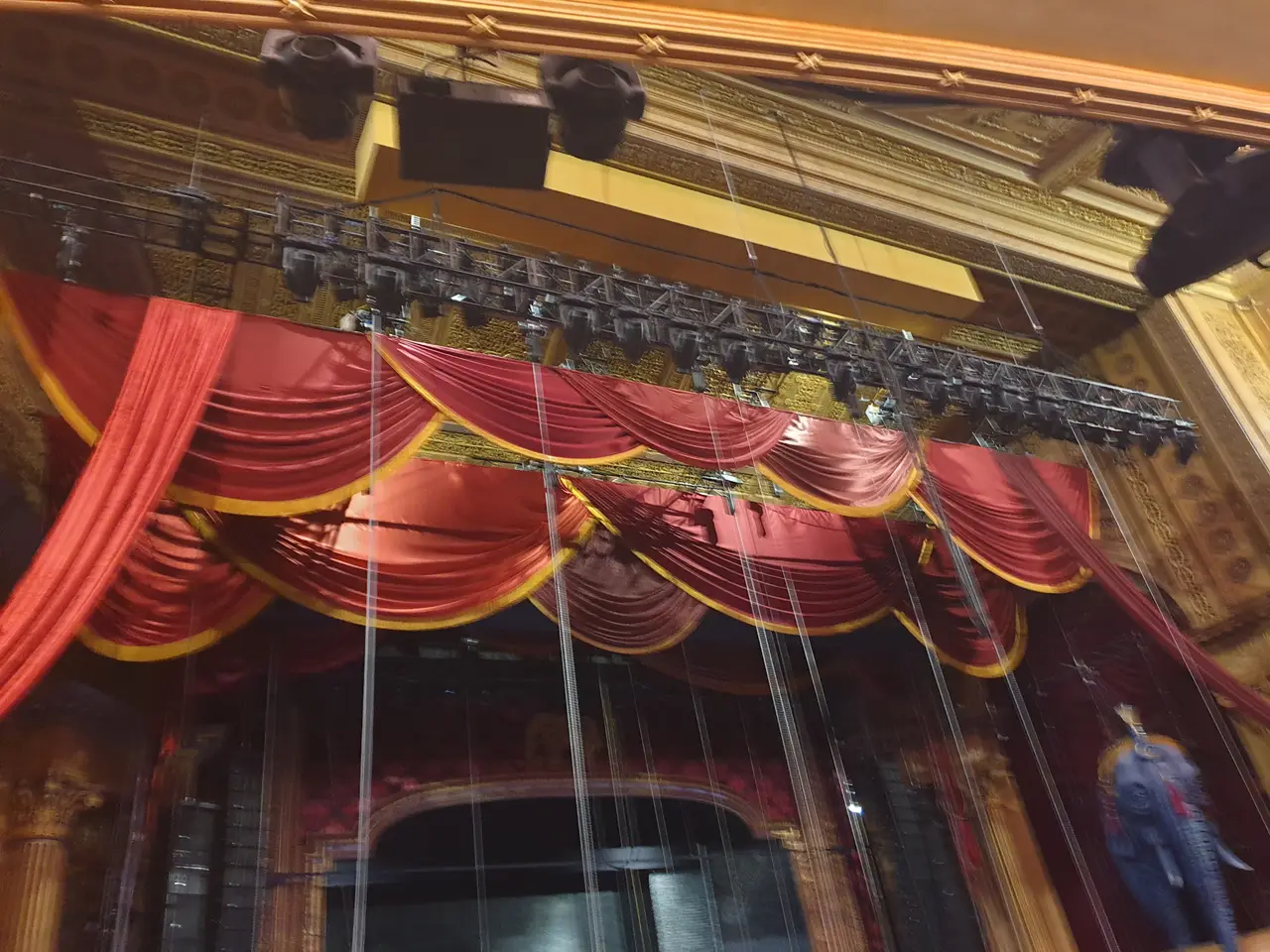 Looking up at red swag drapes with gold trim along lighting truss in an ornate theatre