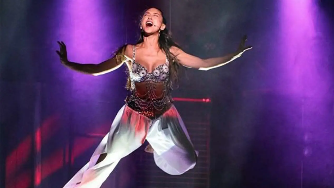 A performer flown mid-air on stage, arms wide, under purple light