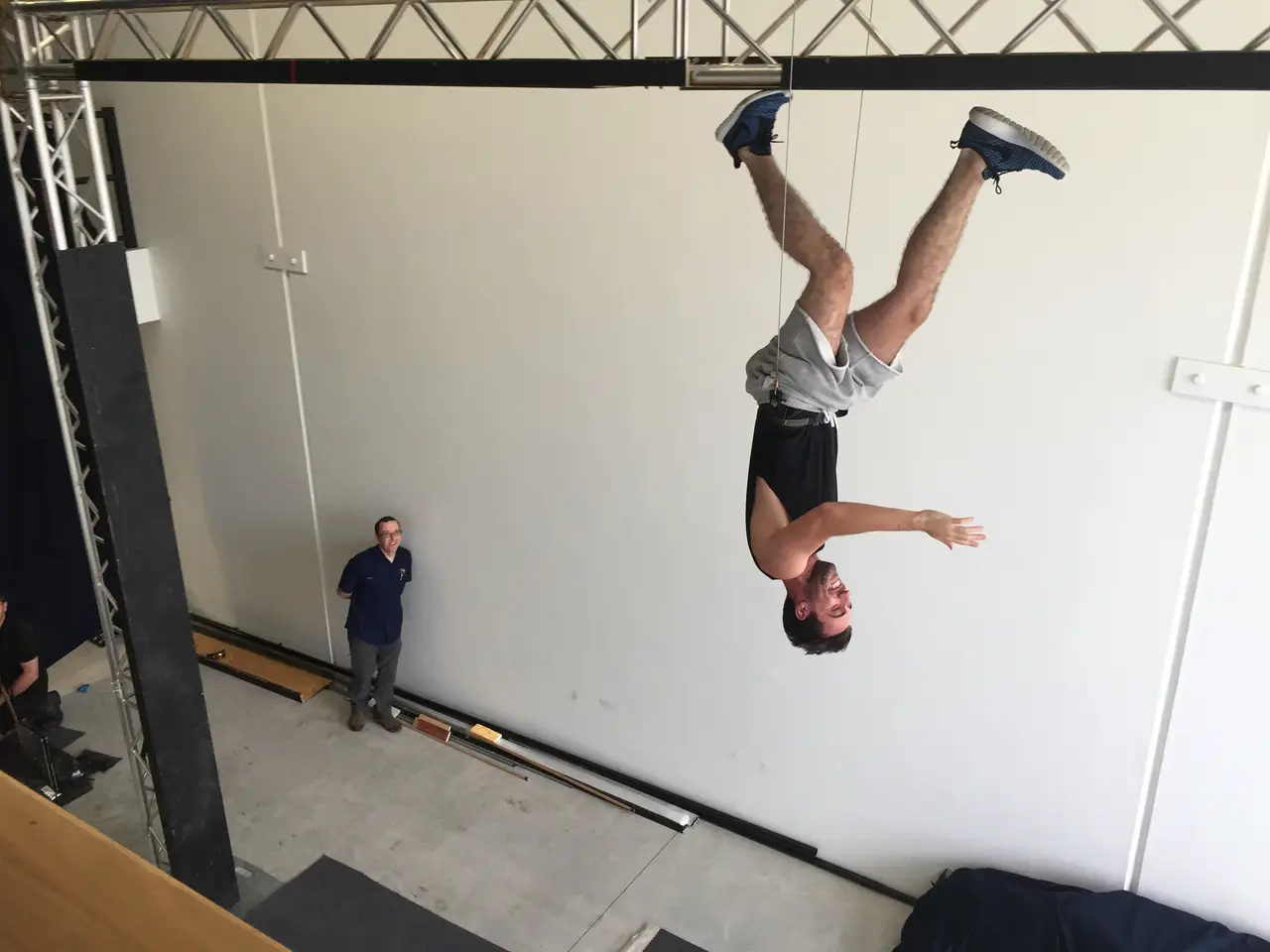 A performer somersaults upside-down on a flying rig under truss while a technician watches, in a bright test space
