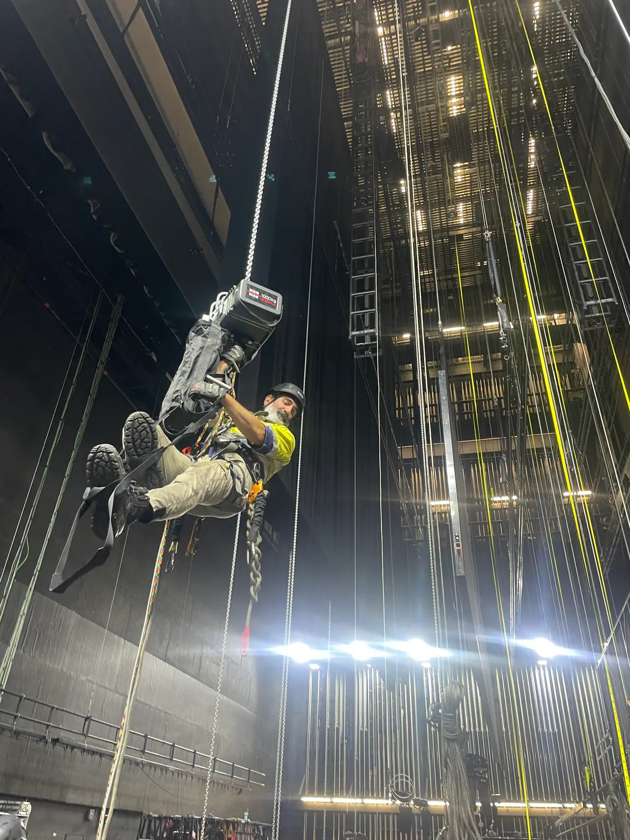 Rigger abseiling on rope beside a motorised hoist in a tall fly tower
