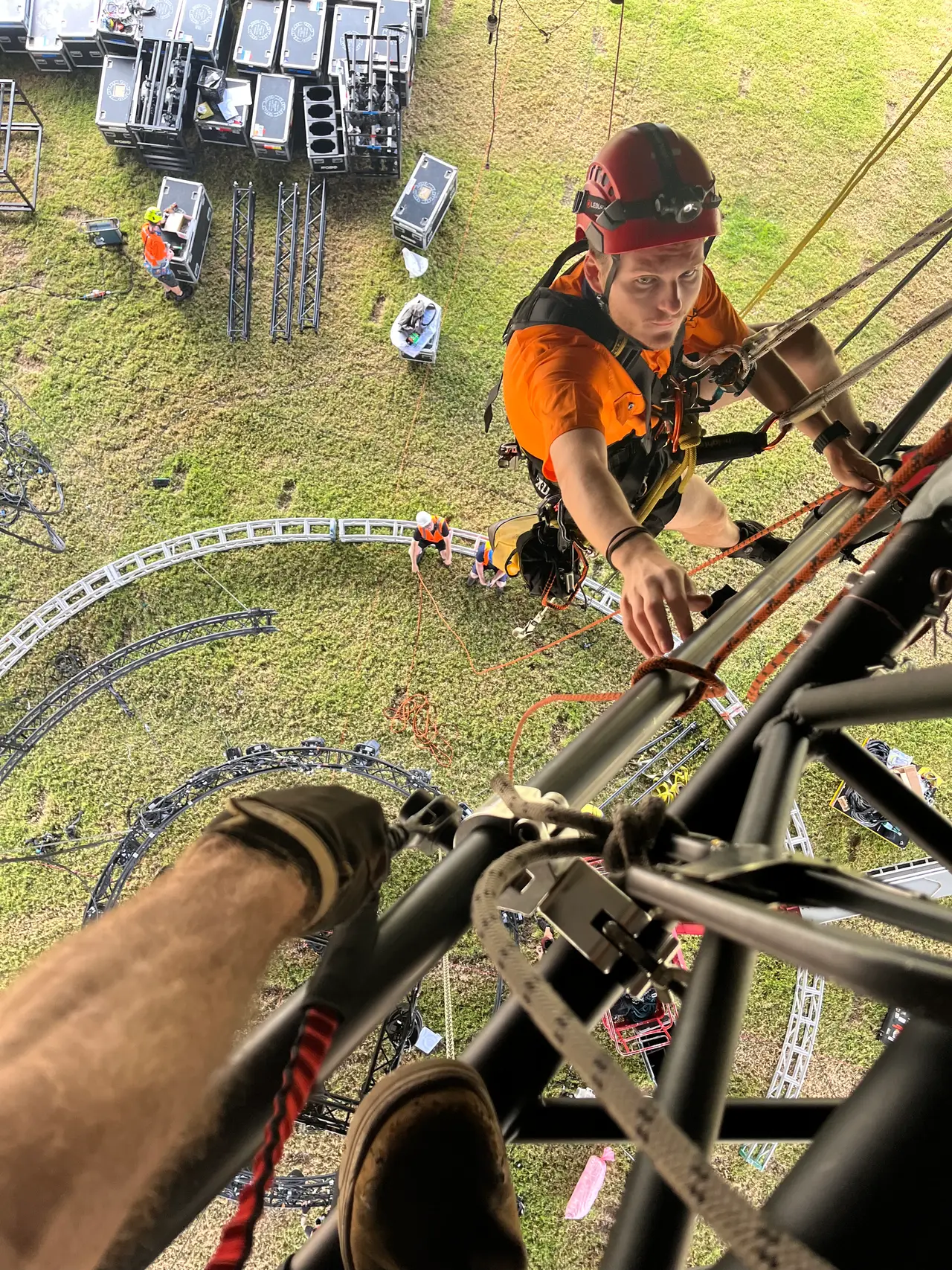 Rigger in harness and helmet descends a truss tower on rope, looking down at gear staged below