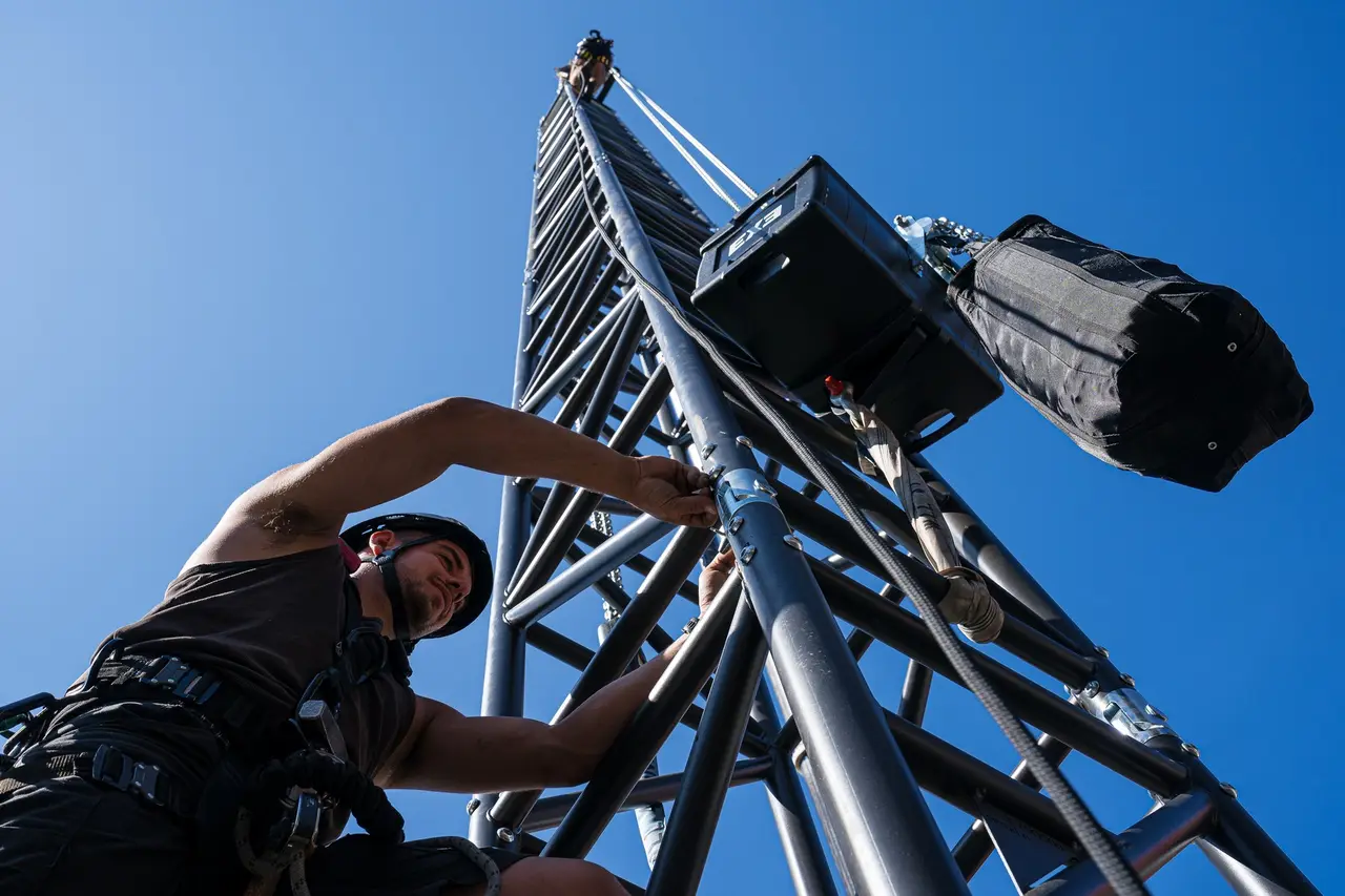 A rigger in helmet and harness climbs a rigging tower with a chain hoist overhead.