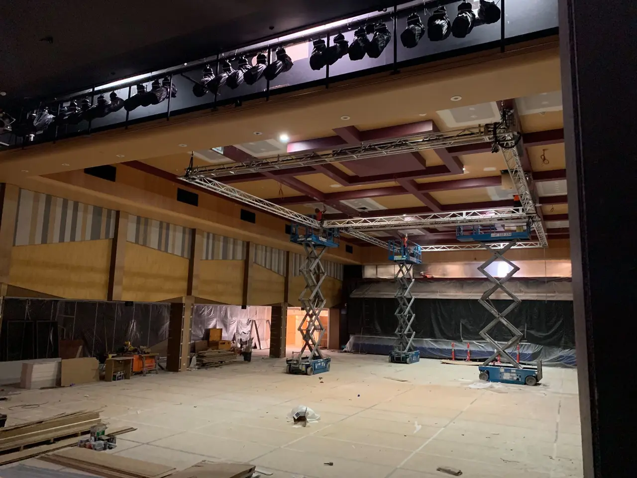 Scissor lifts installing truss over a venue floor still under construction, lights on a gantry above