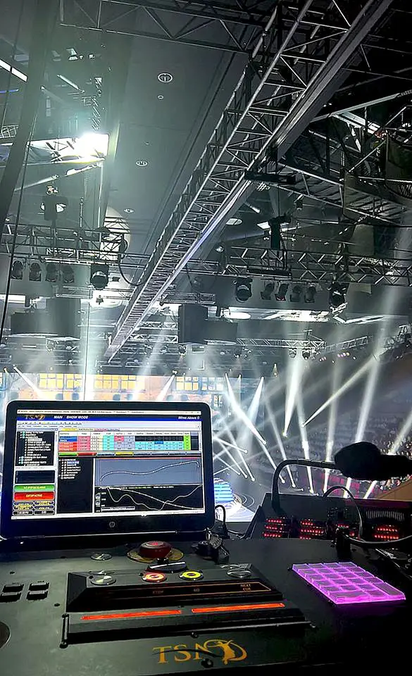 Show control desk and screen in front of arena lighting rig and truss during a concert