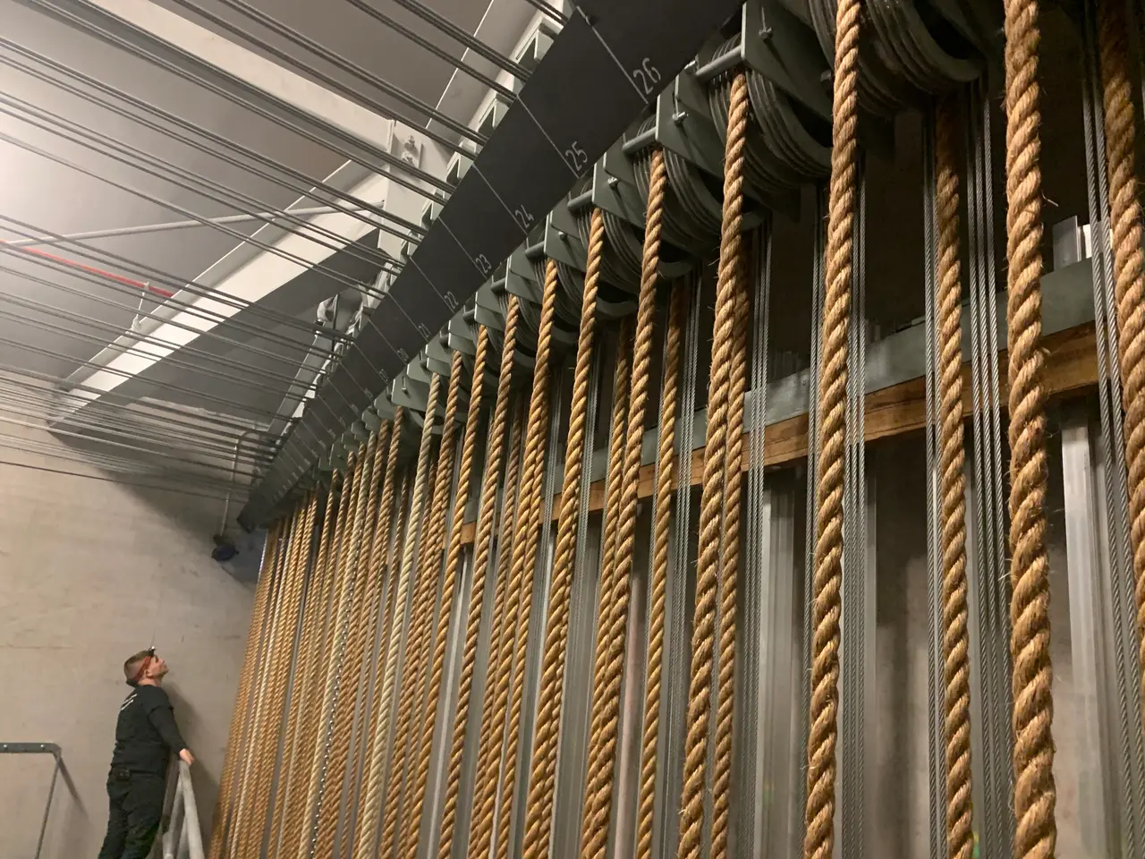 A technician looks up at a bank of counterweight lines and wire ropes running up a fly tower wall.