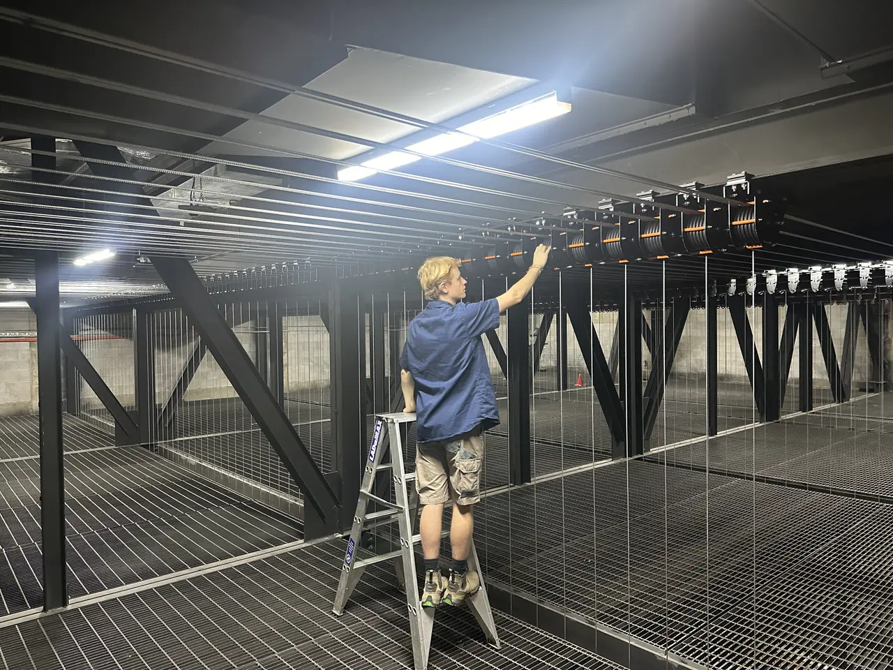 Technician on a stepladder adjusting winches on a fly system with rows of wire lines over a steel grid