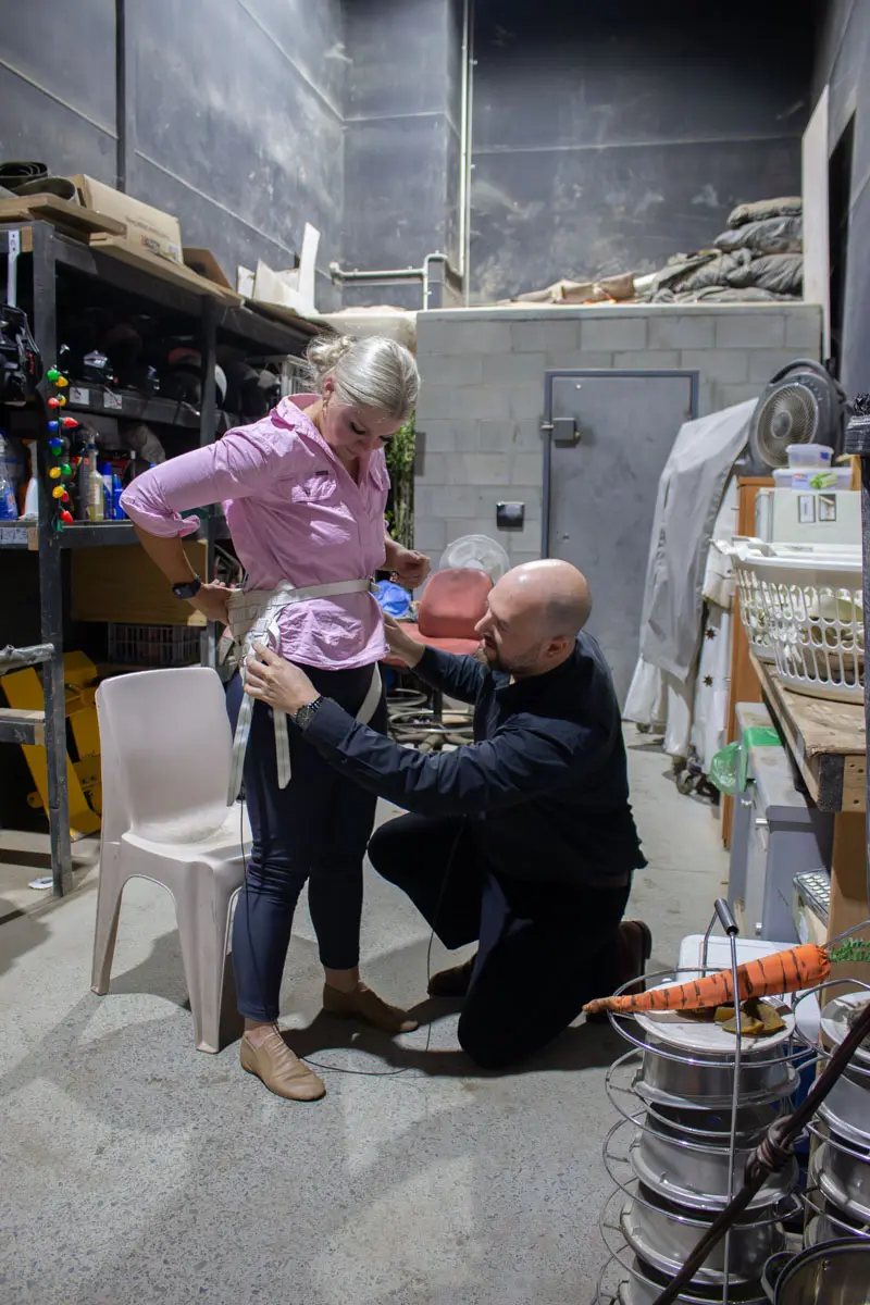 TSA staff member kneels to adjust a flying harness on a woman backstage