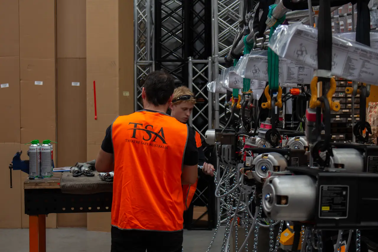 A TSA technician in an orange vest works at the bench on a truss-mounted hoist rig.