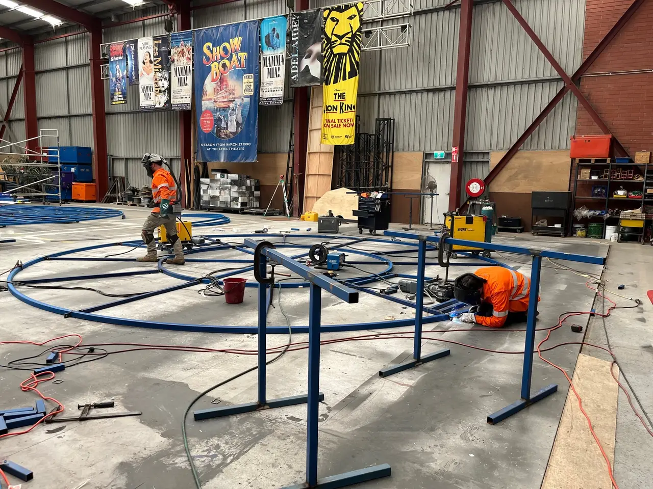 Two riggers welding a circular revolve frame in the workshop, touring show posters behind