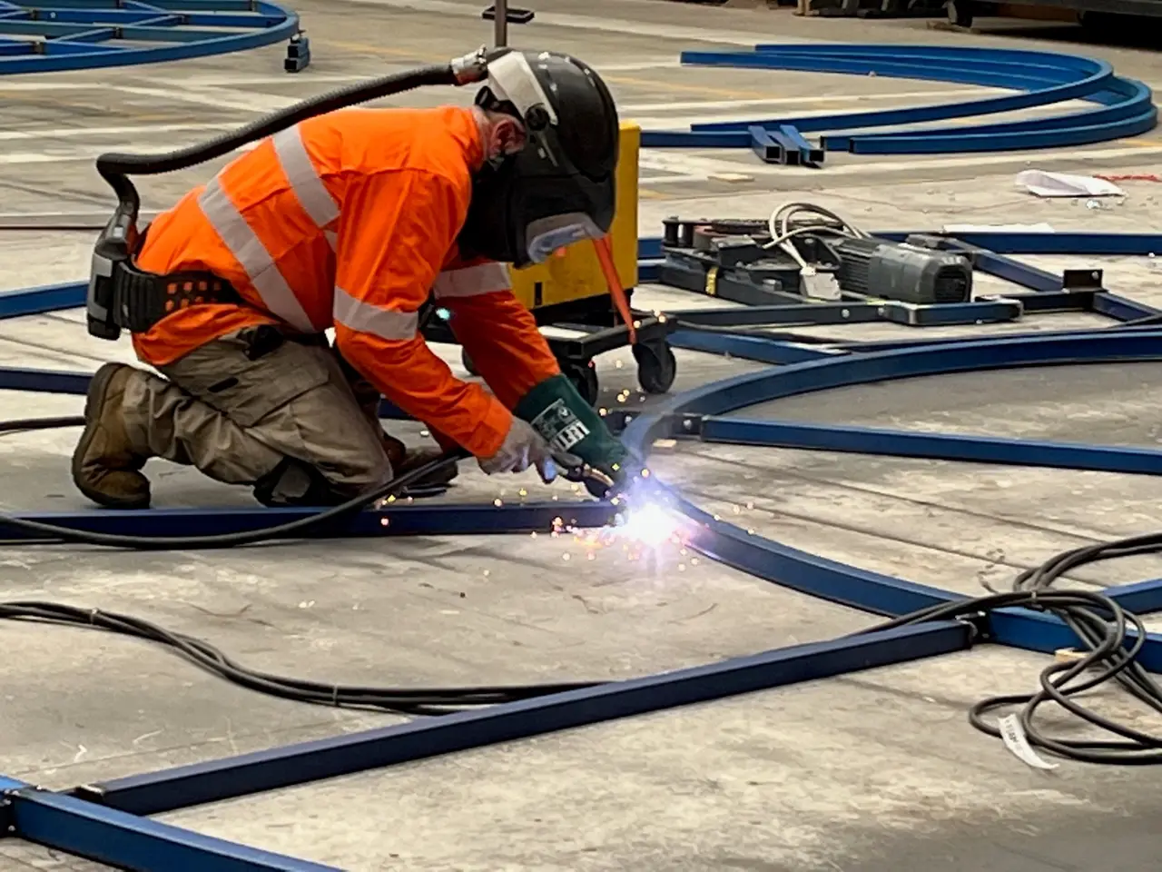 A welder in full PPE fabricates a curved steel revolve track outdoors, sparks flying