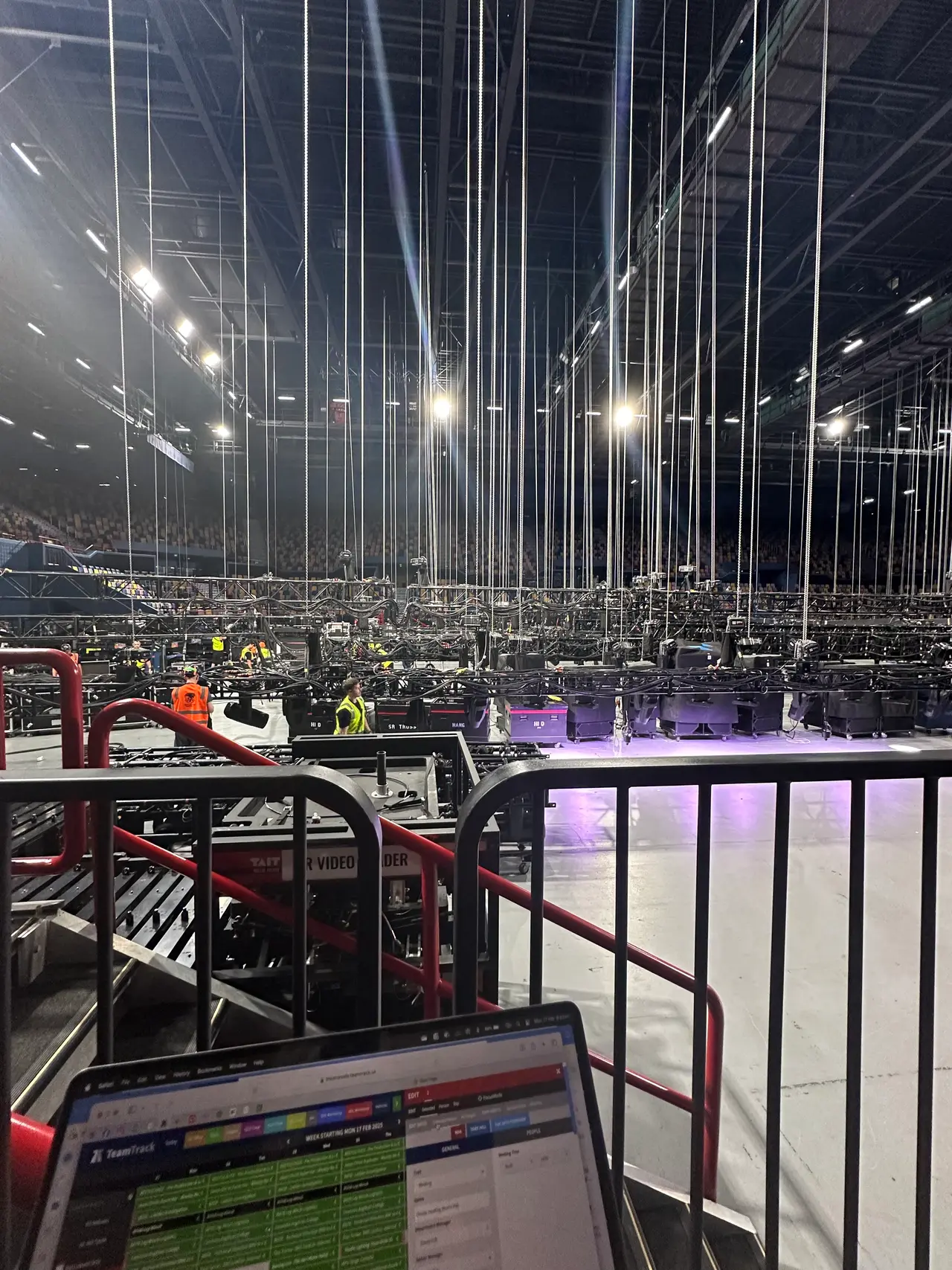 Wide arena rig of chain hoists and truss above the stadium floor with a production laptop in view