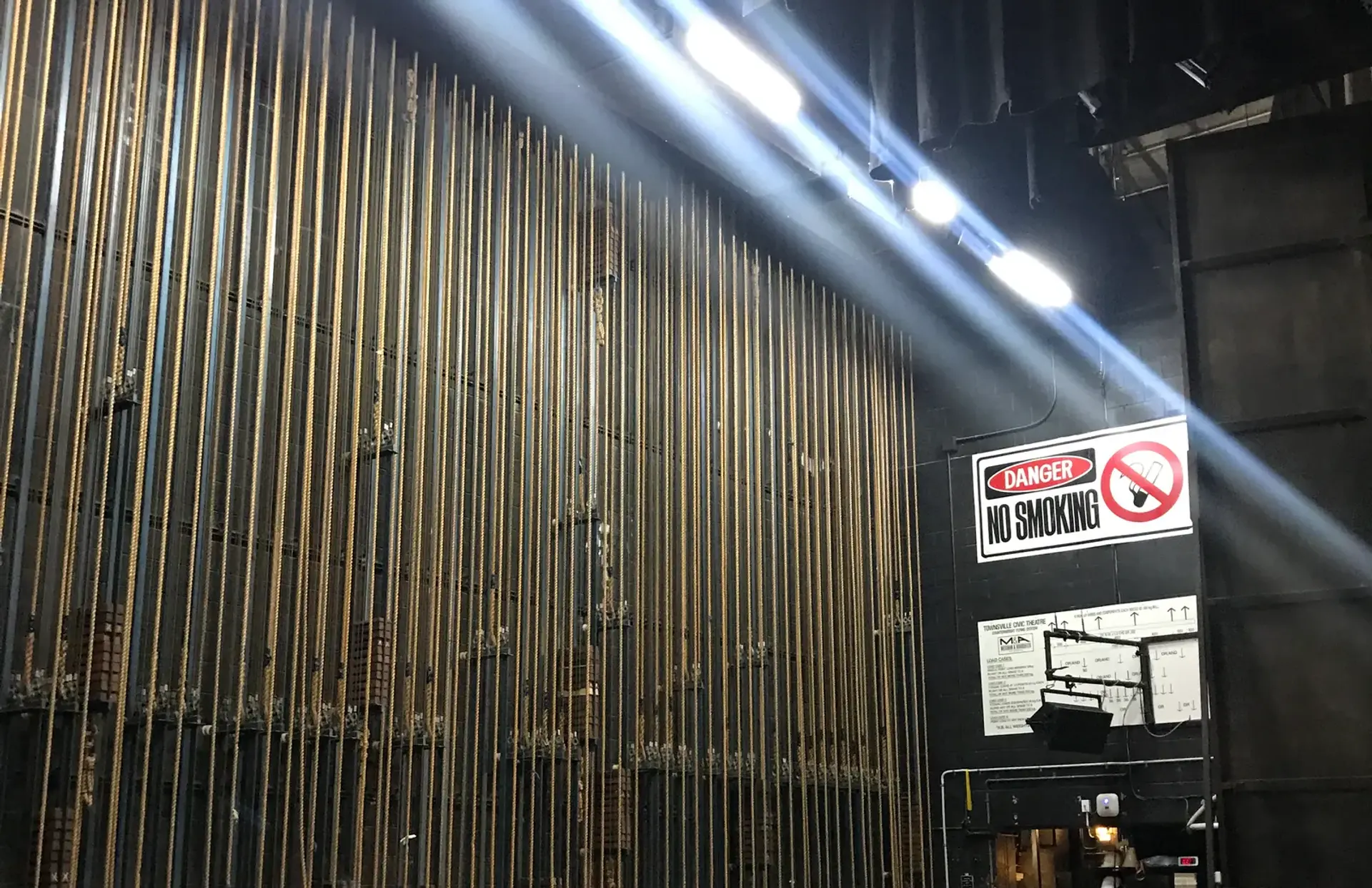 A full wall of counterweight lift lines in a theatre fly tower, lit by stage light