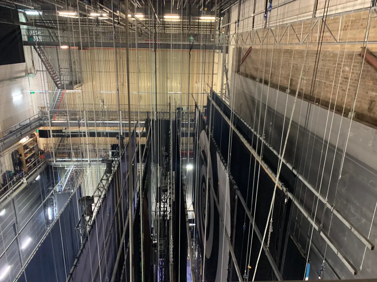 Wire fly lines running down from the grid to black drapes in a theatre fly tower.