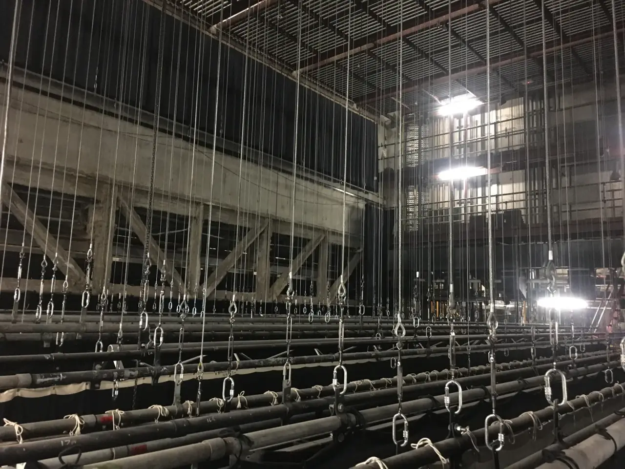 Fly tower grid with hanging wire ropes, shackles and pipe battens above a stage.