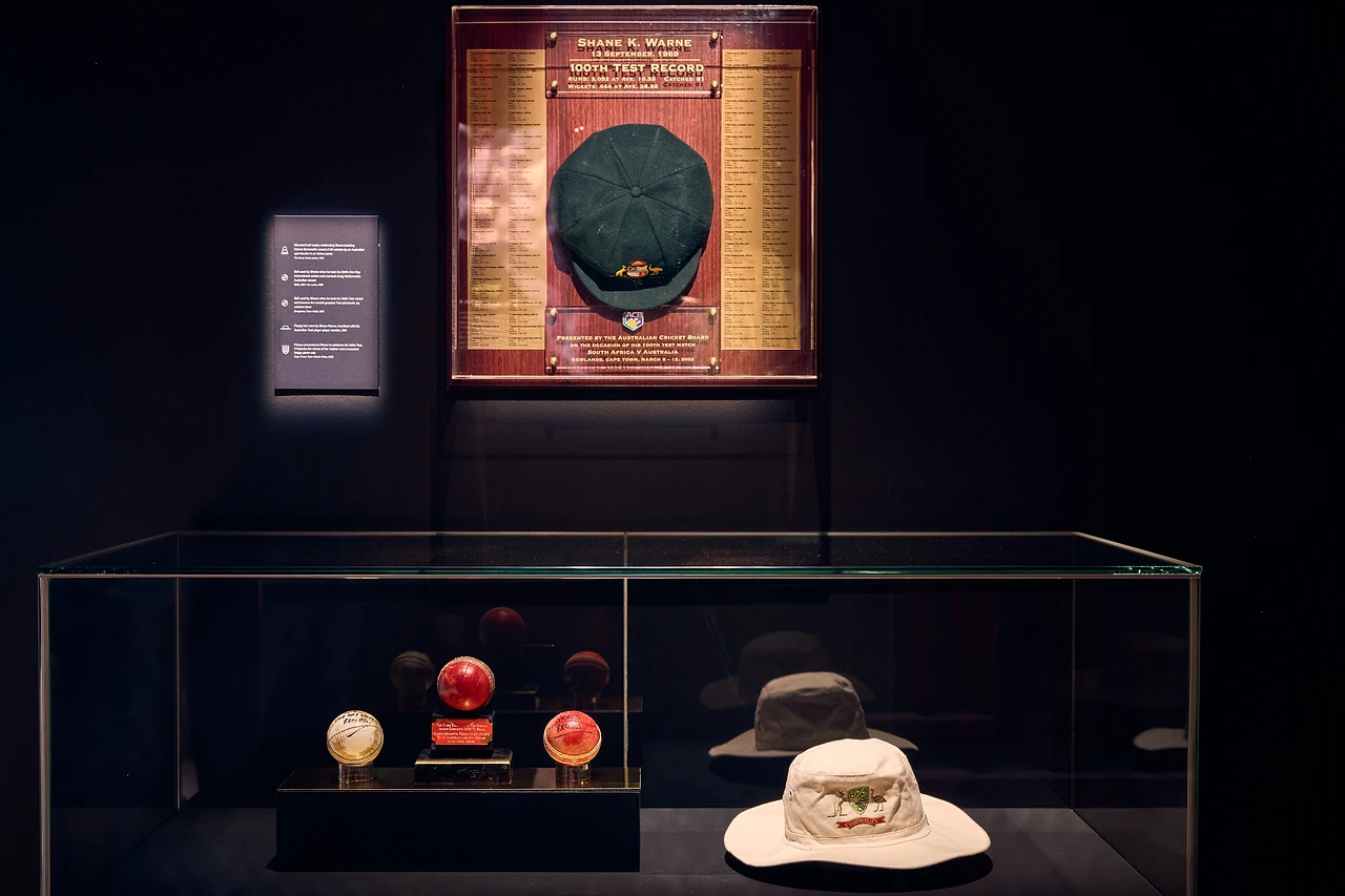 Dark wall showcase with a baggy green cap on a plaque above balls and a sun hat