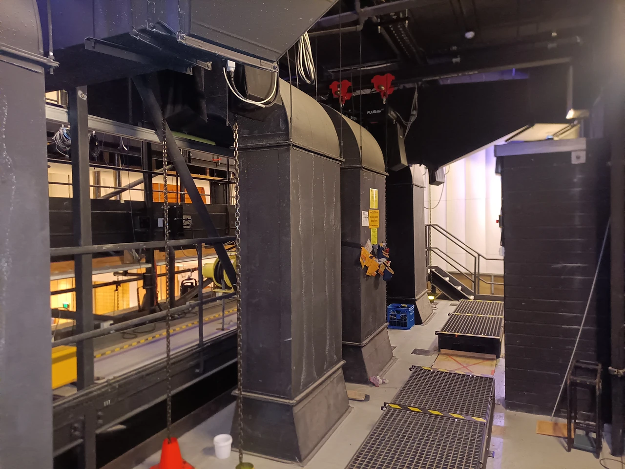 Grated catwalk beside black acoustic ducting above the hall with hoists on trolleys overhead