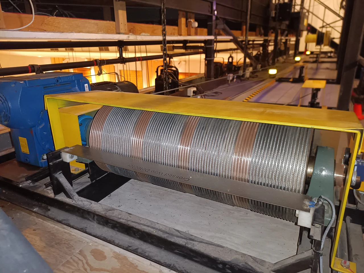 Grooved cable drum inside a yellow guard on a steel gantry above the hall