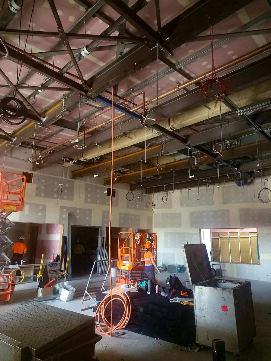 Congested ceiling services above an unfinished studio at ECU City Campus, with a scissor lift on the slab below.