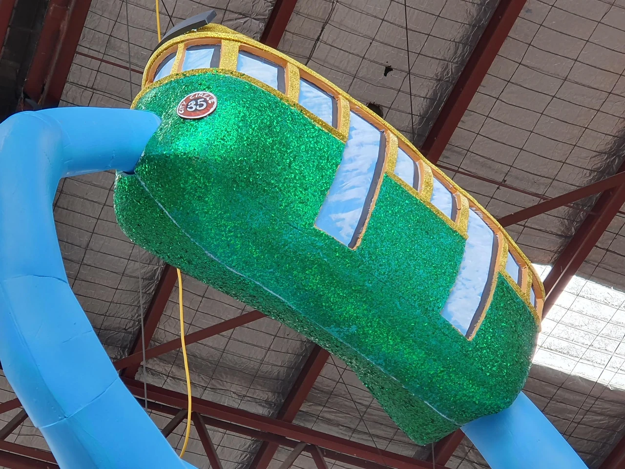 Green glitter tram sculpture hanging from a blue tube in the workshop