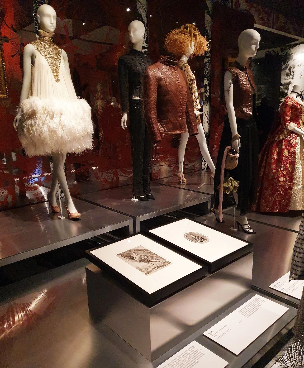 Mannequins in feathered and embroidered garments on a metal-edged plinth with framed drawings