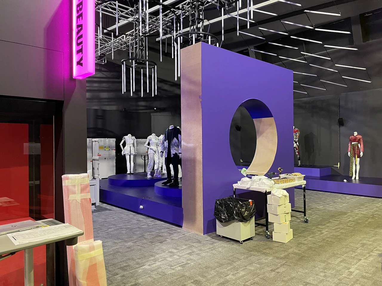 Large purple wall with a round window and neon BEAUTY sign beside a fashion podium