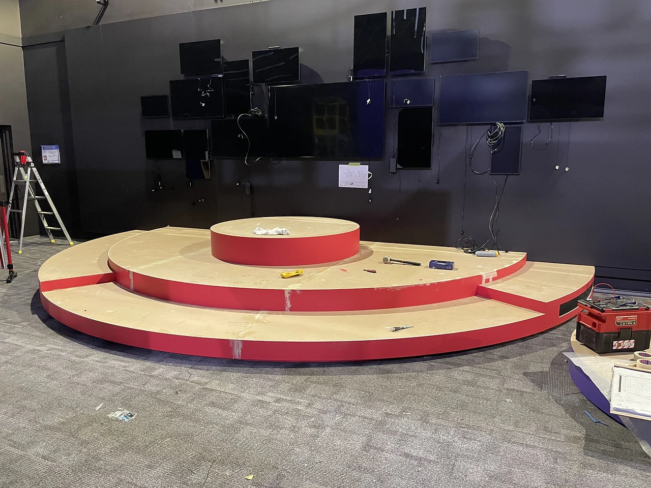 Three-tier red and timber podium under construction beneath a wall of screens