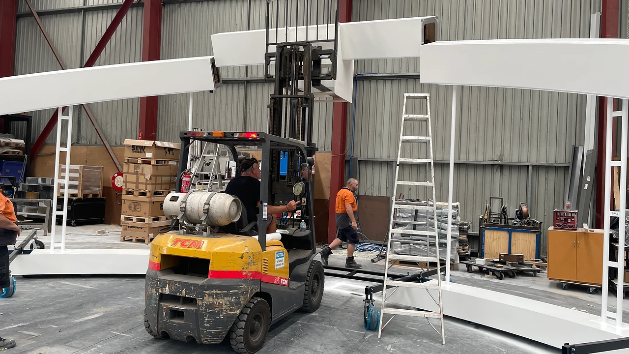 Forklift lifting an arch segment into place above a white ring base in the workshop