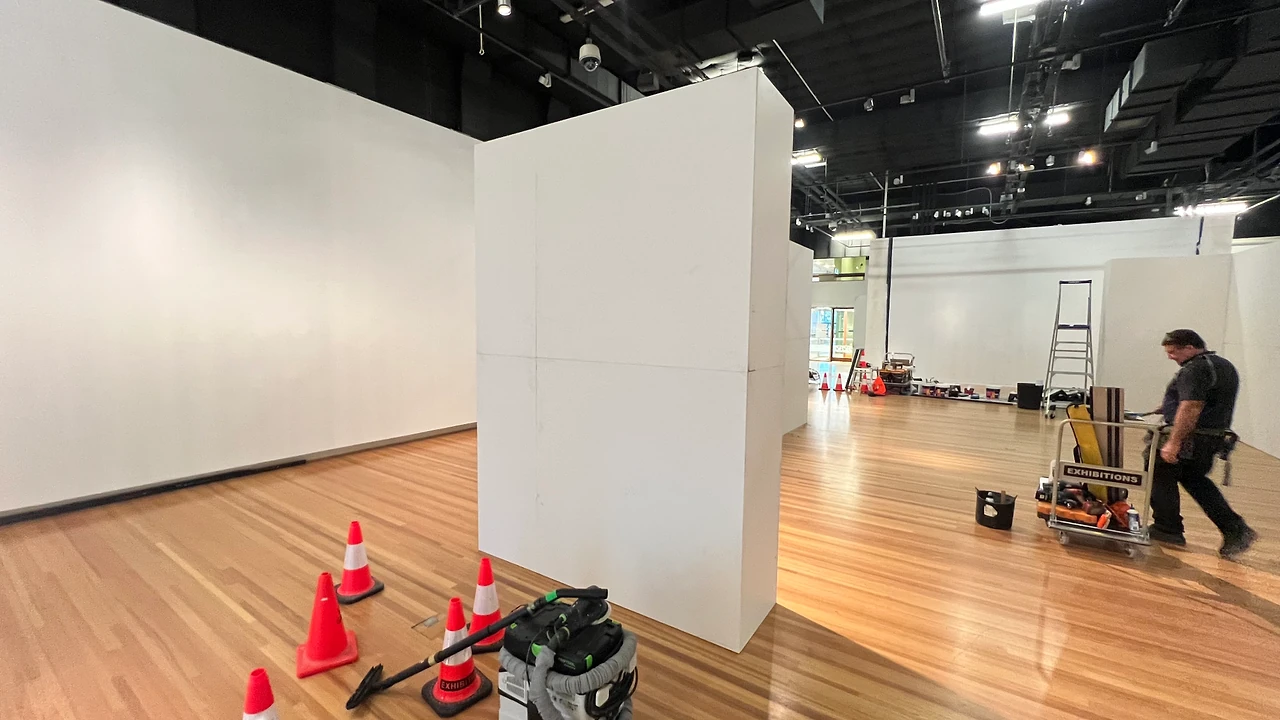 TSA crew member wheeling a tool cart past the freestanding wall panel, with cones and a dust extractor on the timber floor. Design: Tim Blaikie, TSA. Photo: TSA.