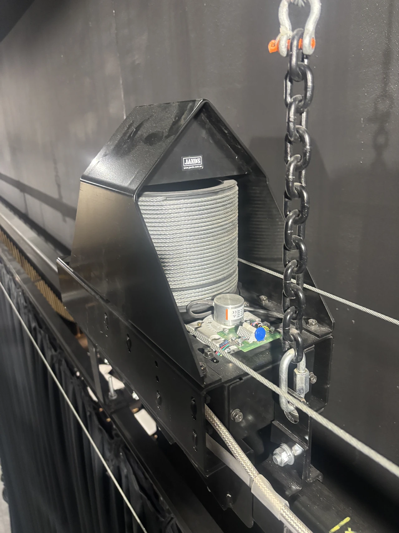 Black motorised hoist housing with wire drum and control board hung on a bar