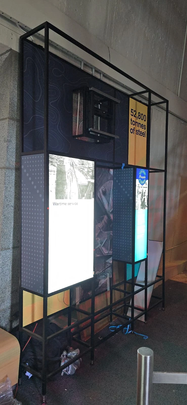 Black steel display frame with backlit panels beside a timber bench and window