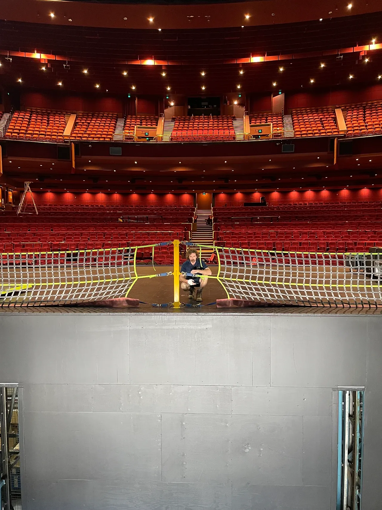 A TSA crew member rigging a safety net across the stage edge on the Wicked tour, the empty auditorium beyond.