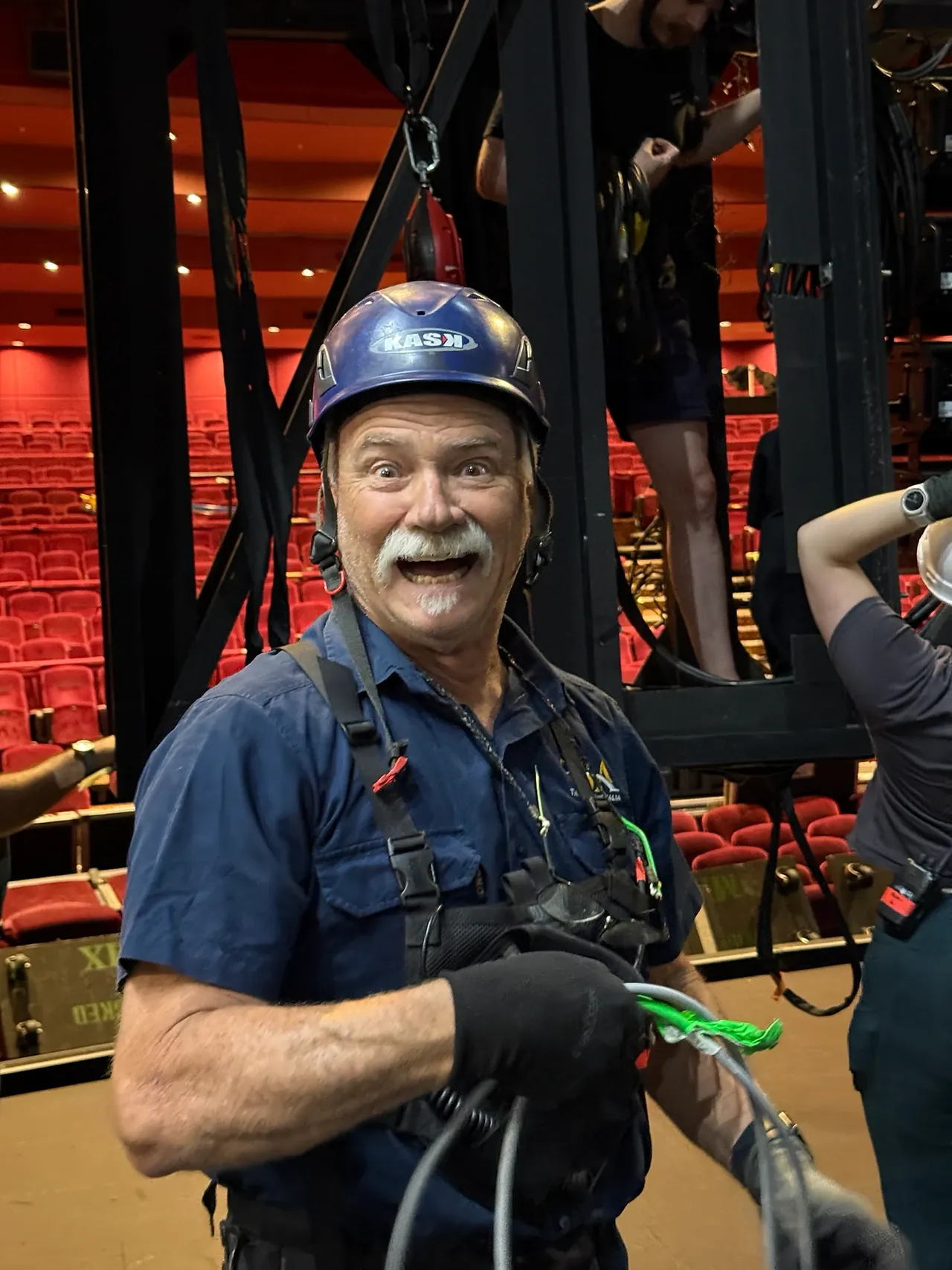 A TSA rigger in full harness and helmet on the Wicked tour, auditorium seating behind.