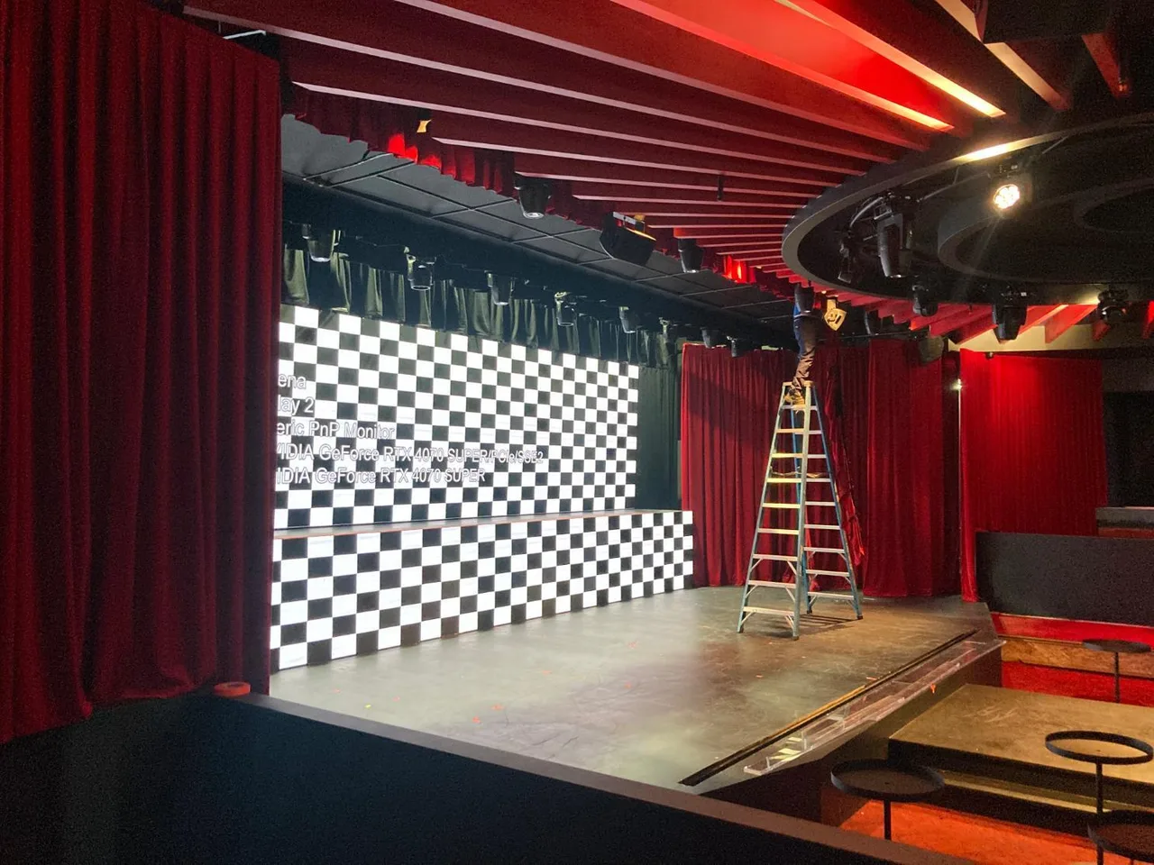 A technician on a ladder rigging red velvet drapes beside an LED screen on a small venue stage