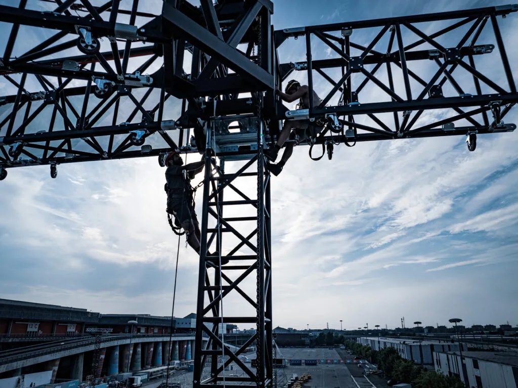 Two riggers climbing a ground-support truss tower and grid above an outdoor venue
